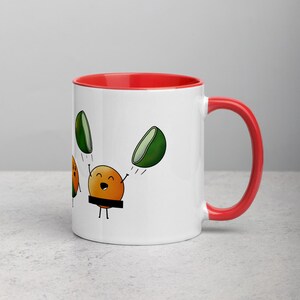 May include: White ceramic mug with a red rim and handle. The mug features a cartoon illustration of a smiling orange with a black belt and two green lime halves flying above it.