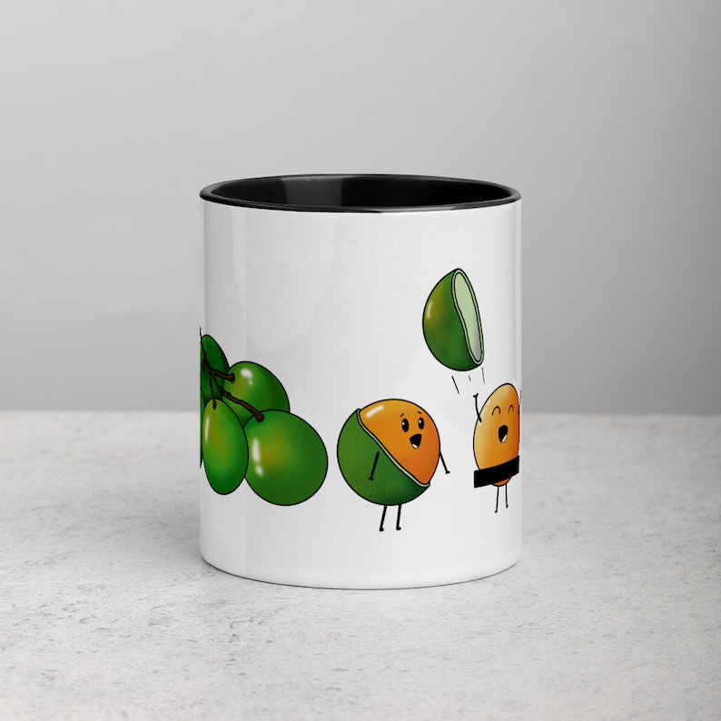 May include: White ceramic mug with a black rim. The mug features a cartoon illustration of green limes and a lime with a face throwing another lime with a face. The lime with a face is wearing a black band around its middle.