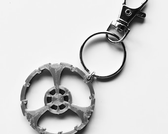 Puede incluir: Un llavero plateado con un diseño circular de tres puntas y un anillo partido plateado unido a un cierre de langosta plateado.
