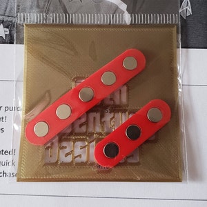 Puede incluir: Dos imanes rectangulares rojos con imanes circulares negros en cada uno. Los imanes est&aacute;n sobre un fondo dorado con el texto "Senti Designs" impreso en &eacute;l.