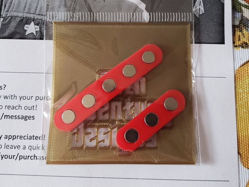 Puede incluir: Dos imanes rectangulares rojos con imanes circulares negros en cada uno. Los imanes est&aacute;n sobre un fondo dorado con el texto "Senti Designs" impreso en &eacute;l.