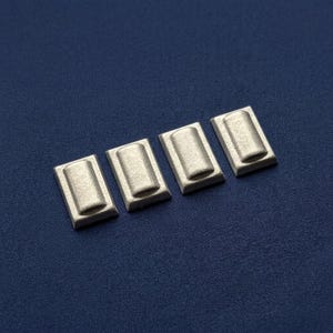 Peut inclure: Quatre petits objets rectangulaires en métal argenté avec une section centrale arrondie en relief. Les objets sont disposés en rangée sur une surface bleu foncé. Le métal a un aspect légèrement texturé.