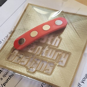 Puede incluir: Una placa decorativa de metal rojo y dorado con la palabra "Century" en relieve. La placa est&aacute; sobre una superficie blanca. Una tira magn&eacute;tica roja y dorada est&aacute; encima de la placa.