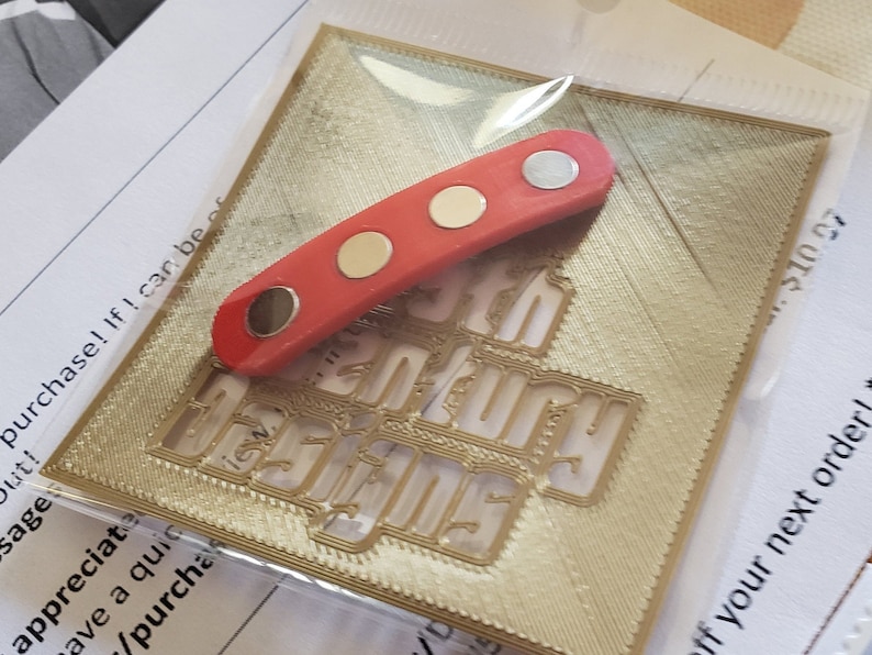 Puede incluir: Una placa decorativa de metal rojo y dorado con la palabra "Century" en relieve. La placa est&aacute; sobre una superficie blanca. Una tira magn&eacute;tica roja y dorada est&aacute; encima de la placa.