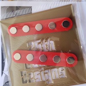 Puede incluir: Dos tiras rojas con cinco botones magn&eacute;ticos plateados cada una. Las tiras est&aacute;n sobre un fondo dorado con un dise&ntilde;o recortado.