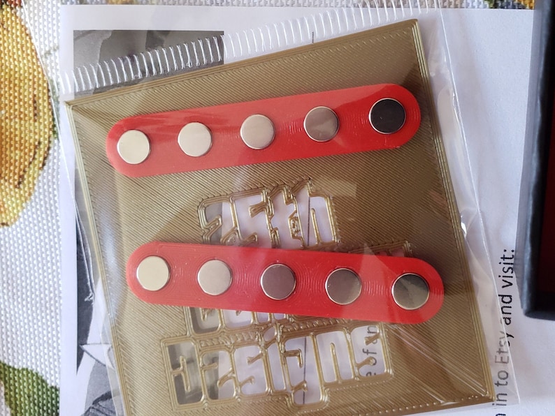 Puede incluir: Dos tiras rojas con cinco botones magn&eacute;ticos plateados cada una. Las tiras est&aacute;n sobre un fondo dorado con un dise&ntilde;o recortado.