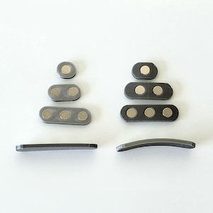 Puede incluir: Un conjunto de clips magn&eacute;ticos grises, cada uno con tres imanes redondos. Los clips est&aacute;n dispuestos en dos pilas, una con cuatro clips y la otra con tres clips. Hay dos clips adicionales, cada uno con una forma curva.