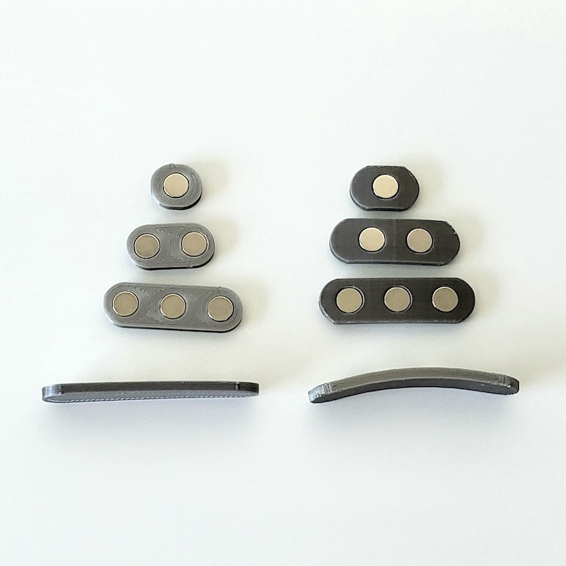 Puede incluir: Un conjunto de clips magn&eacute;ticos grises, cada uno con tres imanes redondos. Los clips est&aacute;n dispuestos en dos pilas, una con cuatro clips y la otra con tres clips. Hay dos clips adicionales, cada uno con una forma curva.