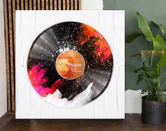 Puede incluir: Un disco de vinilo negro con un diseño abstracto colorido con salpicaduras de pintura rosa, naranja y blanca. El disco está montado sobre un fondo blanco y presenta una etiqueta con el texto "The Miseducation of Lauryn Hill" y un retrato de Lauryn Hill.