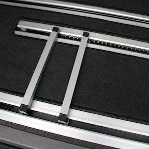 Könnte beinhalten: Nahaufnahme eines Fahrzeuginnenraums mit schwarzem Teppichboden und silbernen Metallschienen. Zwei silberne Metallstützen sind vertikal angeordnet und mit den Schienen verbunden. Das Design deutet auf Autozubehör oder Innenraumkomponenten hin.