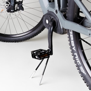 Può includere: Una mountain bike elettrica grigia con un cavalletto nero e un pedale nero. La bici ha un pneumatico nero con lettere bianche che dicono "Derby Nic".