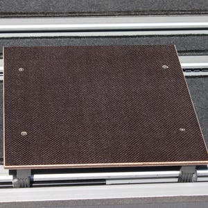 Könnte beinhalten: Eine rechteckige braune Holzplattform mit strukturierter Oberfläche, befestigt mit vier silbernen Schrauben. Die Plattform ist auf einem silbernen Schienensystem mit schwarzen Kunststoffhaltern montiert, das zum Verschieben oder zur Einstellung der Position dient.