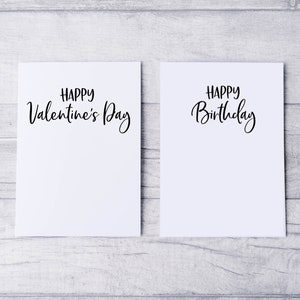 May include: Two blank white cards with black calligraphy. The card on the left says "Happy Valentine's Day". The card on the right says "Happy Birthday".