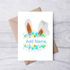 May include: A white greeting card with watercolor bunny ears and a floral design in blue, green, and orange. The text "Add Name" is written in blue. The card is inside a brown envelope.