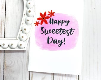 May include: A white greeting card with the text "Happy Sweetest Day!" in black script, set against a pink watercolor wash. Red flower illustrations are in the upper left corner. A white, beaded frame is in the upper left.