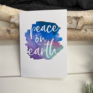 May include: A white card with a watercolor background in shades of blue, purple, and green. The text "peace on earth" is written in white cursive font.