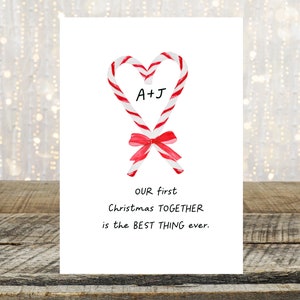 May include: A white card with a red and white candy cane heart design. The candy canes are tied together with a red ribbon. The text "A+J" is inside the heart. The text "OUR first Christmas TOGETHER is the BEST THING ever." is below the heart.