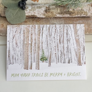 May include: A snowy forest scene with a small evergreen tree in the center. The text "MAY YOUR TRAILS BE MERRY + BRIGHT." is printed at the bottom of the card.