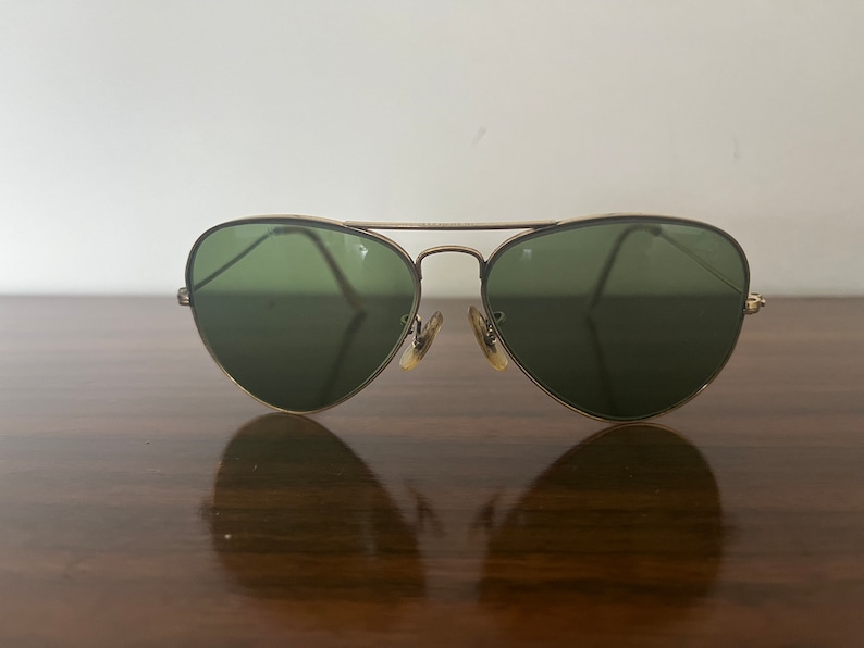 Peut inclure: Lunettes de soleil aviateur dor&eacute;es avec des verres verts. Les lunettes sont pos&eacute;es sur une surface en bois.