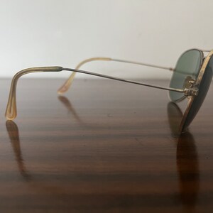 Peut inclure: Lunettes de soleil aviateur dor&eacute;es avec des verres verts et des branches en plastique brun. Les lunettes de soleil reposent sur une surface en bois.