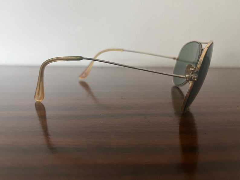 Peut inclure: Lunettes de soleil aviateur dor&eacute;es avec des verres verts et des branches en plastique brun. Les lunettes de soleil reposent sur une surface en bois.