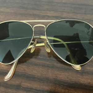 Peut inclure: Lunettes de soleil aviateur dor&eacute;es avec des verres vert fonc&eacute;. Les lunettes sont pos&eacute;es sur une surface en bois.