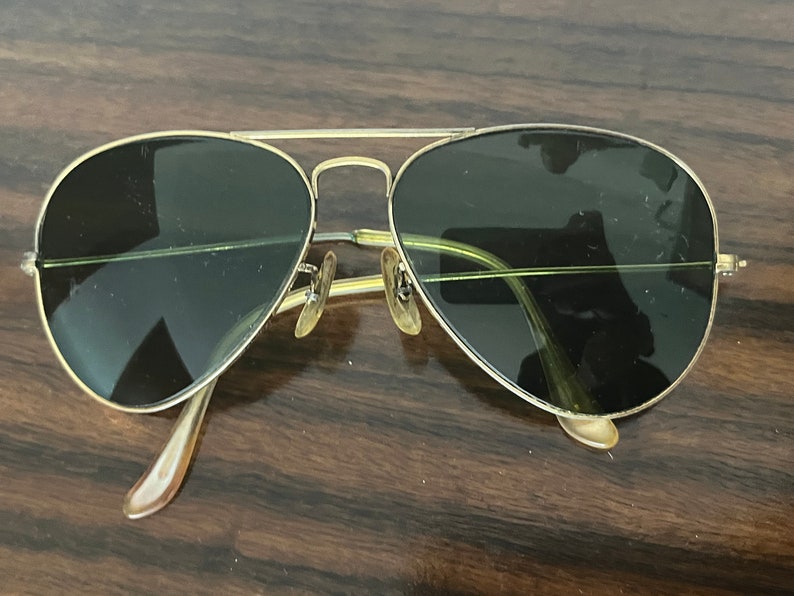 Peut inclure: Lunettes de soleil aviateur dor&eacute;es avec des verres vert fonc&eacute;. Les lunettes sont pos&eacute;es sur une surface en bois.
