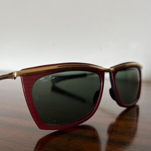 Op de afbeelding: Rode en gouden Ray-Ban zonnebril met een uniek, hoekig montuur. De zonnebril heeft groene glazen.