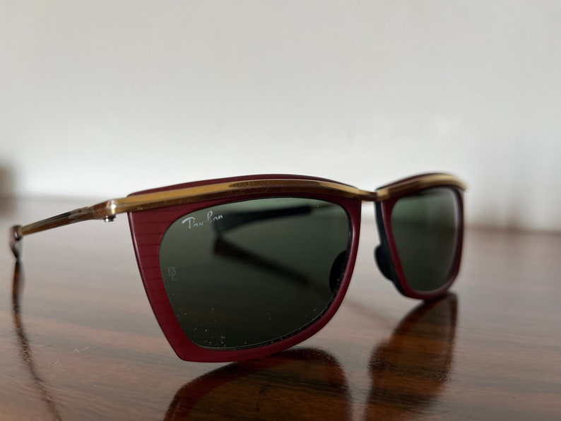 Op de afbeelding: Rode en gouden Ray-Ban zonnebril met een uniek, hoekig montuur. De zonnebril heeft groene glazen.
