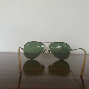 Peut inclure: Une paire de lunettes de soleil aviateur dor&eacute;es avec des verres verts. Les lunettes sont pos&eacute;es sur une surface en bois, les branches &eacute;tant d&eacute;ploy&eacute;es sur les c&ocirc;t&eacute;s.