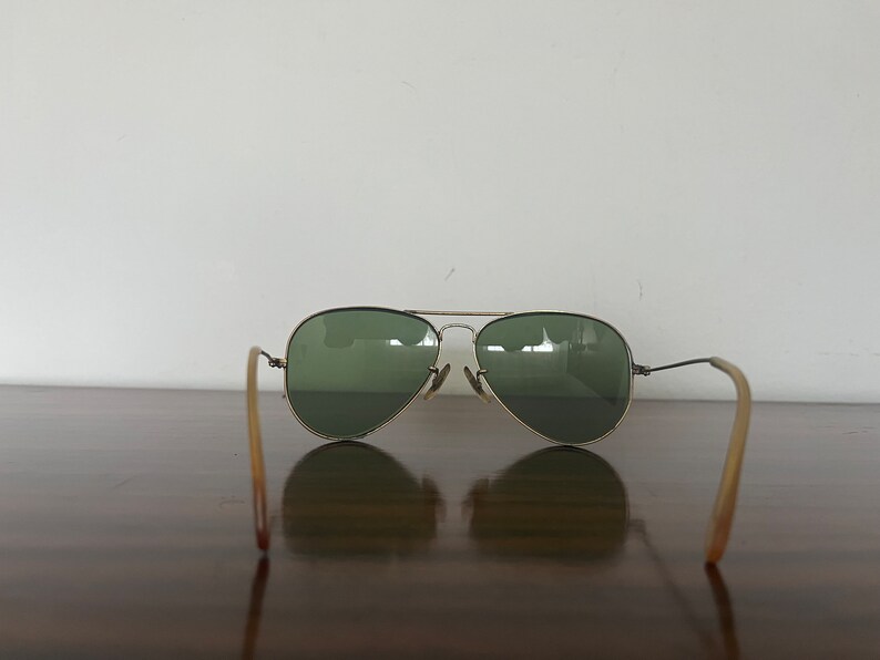 Peut inclure: Une paire de lunettes de soleil aviateur dor&eacute;es avec des verres verts. Les lunettes sont pos&eacute;es sur une surface en bois, les branches &eacute;tant d&eacute;ploy&eacute;es sur les c&ocirc;t&eacute;s.