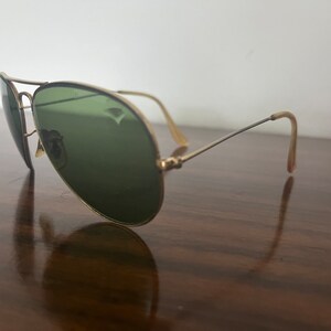Peut inclure: Lunettes de soleil aviateur dor&eacute;es avec des verres verts. Les lunettes sont pos&eacute;es sur une surface en bois.