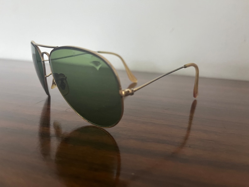 Peut inclure: Lunettes de soleil aviateur dor&eacute;es avec des verres verts. Les lunettes sont pos&eacute;es sur une surface en bois.