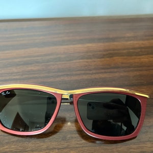 Op de afbeelding: Rode zonnebril met een gouden rand en donkere glazen. De merknaam "Ray-Ban" is zichtbaar op de linkerlens.