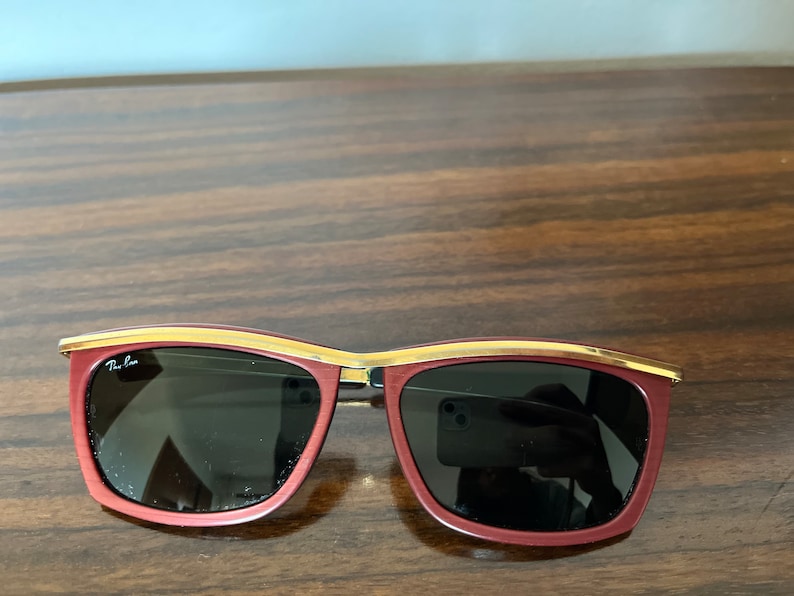 Op de afbeelding: Rode zonnebril met een gouden rand en donkere glazen. De merknaam "Ray-Ban" is zichtbaar op de linkerlens.