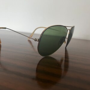 Peut inclure: Une paire de lunettes de soleil aviateur dor&eacute;es avec des verres verts. Les lunettes sont pos&eacute;es sur une surface en bois.