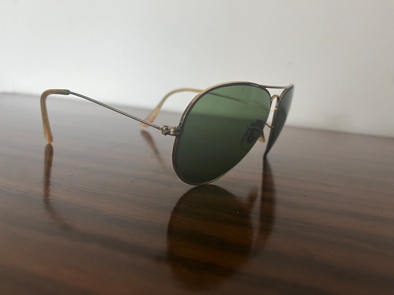 Peut inclure: Une paire de lunettes de soleil aviateur dor&eacute;es avec des verres verts. Les lunettes sont pos&eacute;es sur une surface en bois.