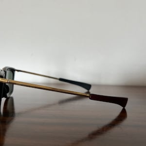 Op de afbeelding: Een zonnebril met een gouden montuur, donkere glazen en bordeauxrode accenten. De zonnebril ligt op een houten oppervlak.