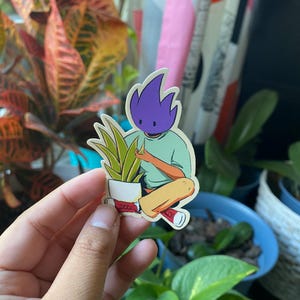 May include: A sticker depicting a cartoon figure with a purple head, light green shirt, and yellow trousers, caring for a potted plant. The figure is seated, wearing red shoes. The sticker is held in a hand, with plants in the background.