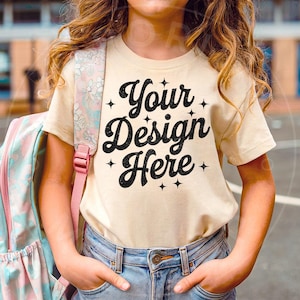 May include: A young person wearing a beige t-shirt with the text "Your Design Here" in black lettering. The shirt has a distressed look and is decorated with small stars. The person is wearing blue jeans and has a pink and blue backpack.