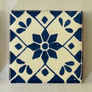May include: A white ceramic tile with a blue and white geometric pattern. The tile features a central flower design with eight points and a smaller circle in the center. The tile is surrounded by a repeating pattern of diamonds and teardrop shapes.