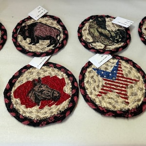 May include: A collection of eight oval-shaped coasters with braided jute centers and black and red borders. Each coaster features a different animal or patriotic design, including a moose, buffalo, bird, bear, horse, and a pumpkin.