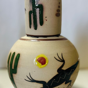 May include: A ceramic bottle with a removable top, decorated with a southwestern design. The bottle features a green cactus, a black lizard, and a yellow sun with red accents.