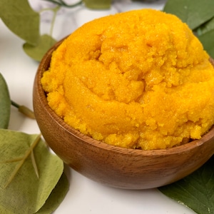 May include: A wooden bowl filled with a bright yellow, grainy scrub. The scrub is likely a body scrub or face scrub.