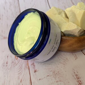 May include: A blue jar of shea butter cream with the text "Glow & Glow" and "brighter, tighter, healthier skin" on the label. The jar is sitting on a white wooden surface next to a wooden bowl filled with raw shea butter.