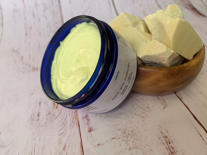 May include: A blue jar of shea butter cream with the text "Glow & Glow" and "brighter, tighter, healthier skin" on the label. The jar is sitting on a white wooden surface next to a wooden bowl filled with raw shea butter.