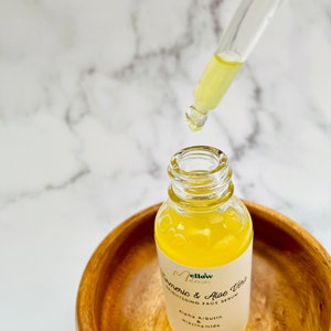 Puede incluir: Una botella de vidrio transparente con un l&iacute;quido amarillo en su interior. La botella est&aacute; etiquetada con "Mellow Elixir Turmeric & Aloe Vera Brightening Face Serum" con los ingredientes "Alpha Arbutin & Niacinamide" enumerados debajo. La botella est&aacute; sentada en un taz&oacute;n de madera.