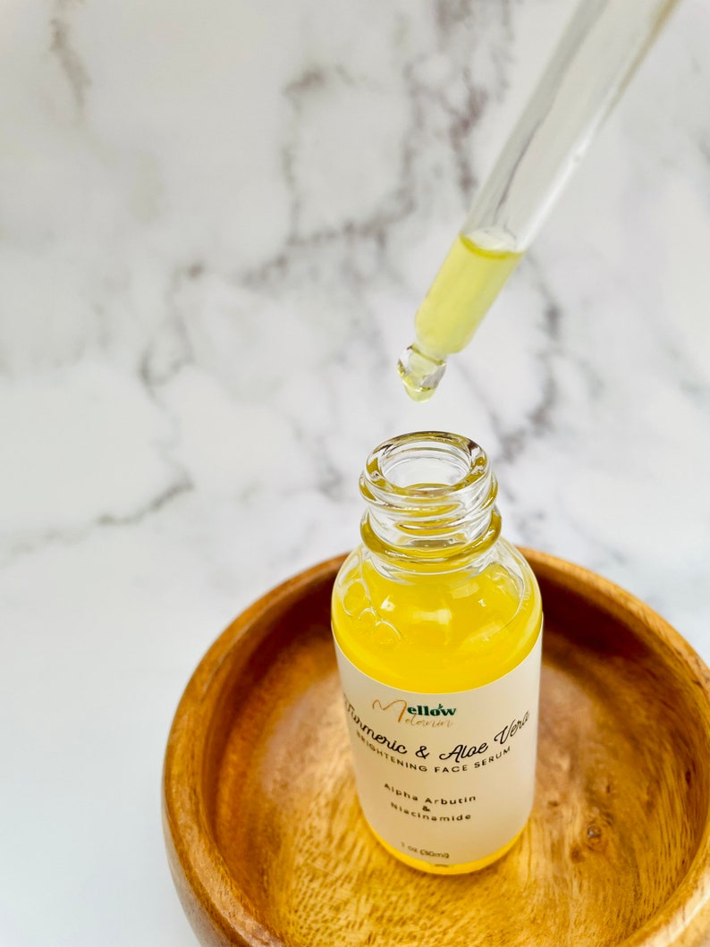Puede incluir: Una botella de vidrio transparente con un l&iacute;quido amarillo en su interior. La botella est&aacute; etiquetada con "Mellow Elixir Turmeric & Aloe Vera Brightening Face Serum" con los ingredientes "Alpha Arbutin & Niacinamide" enumerados debajo. La botella est&aacute; sentada en un taz&oacute;n de madera.
