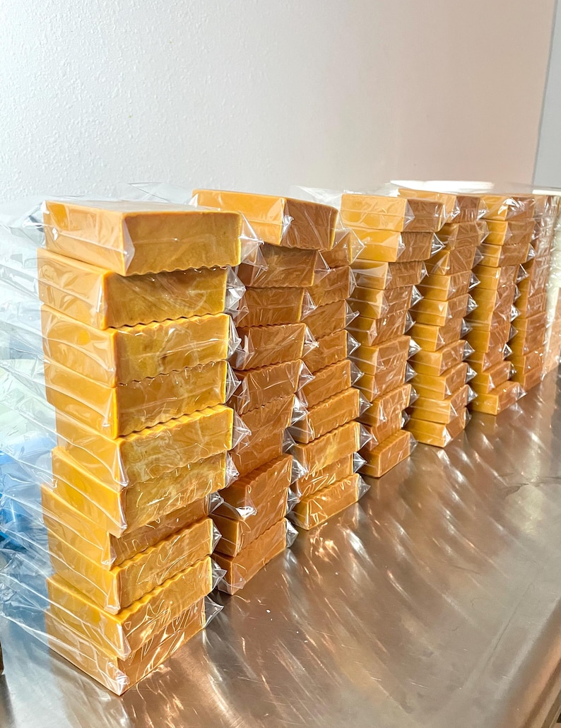 Pu&ograve; includere: Una pila di saponette di colore giallo dorato, avvolte singolarmente. Il sapone &egrave; impilato in file su una superficie argentata.