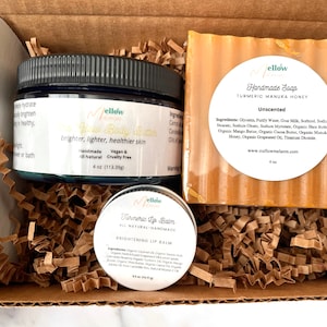 May include: A gift box filled with brown crinkle paper contains three beauty products: a jar of turmeric body butter, a bar of turmeric manuka honey soap, and a small tin of turmeric lip balm. All products are labeled "Mellow" and are handmade and all-natural.